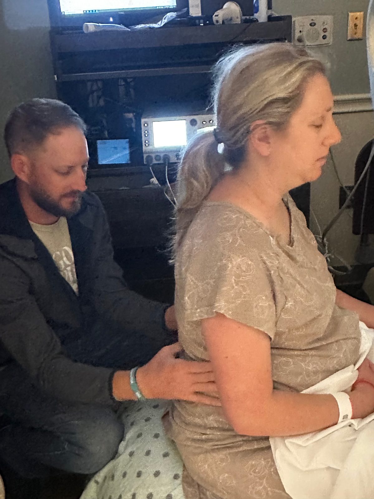 A mom in labor sitting on the edge of a hospital bed, eyes closed, while her husband presses on her lower back