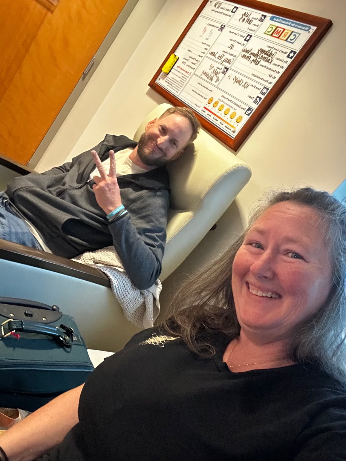 Kristie smiling in a hospital room while Sarah Jane’s dad gives a peace sign from the recliner behind her