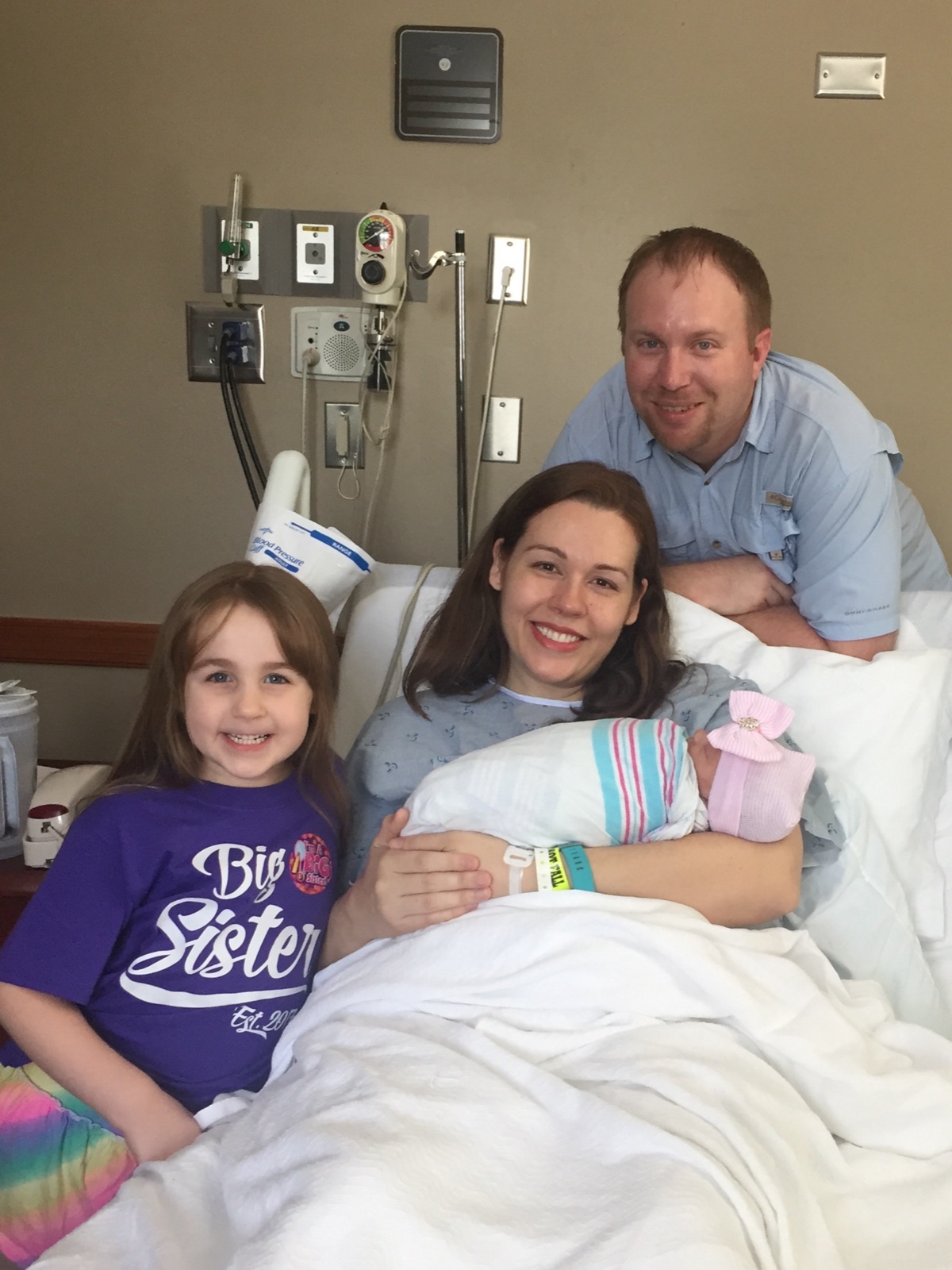 A smiling family in a hospital room: mom holding her newborn, dad behind, and a big sister in a “Big Sister” shirt