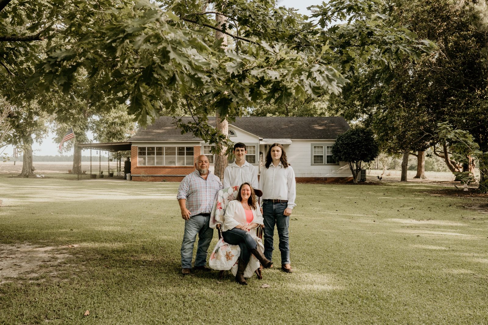 Kristie in a rocking chair draped with a quilt, her husband and two boys standing beside her under an oak tree, a farmhouse behind them