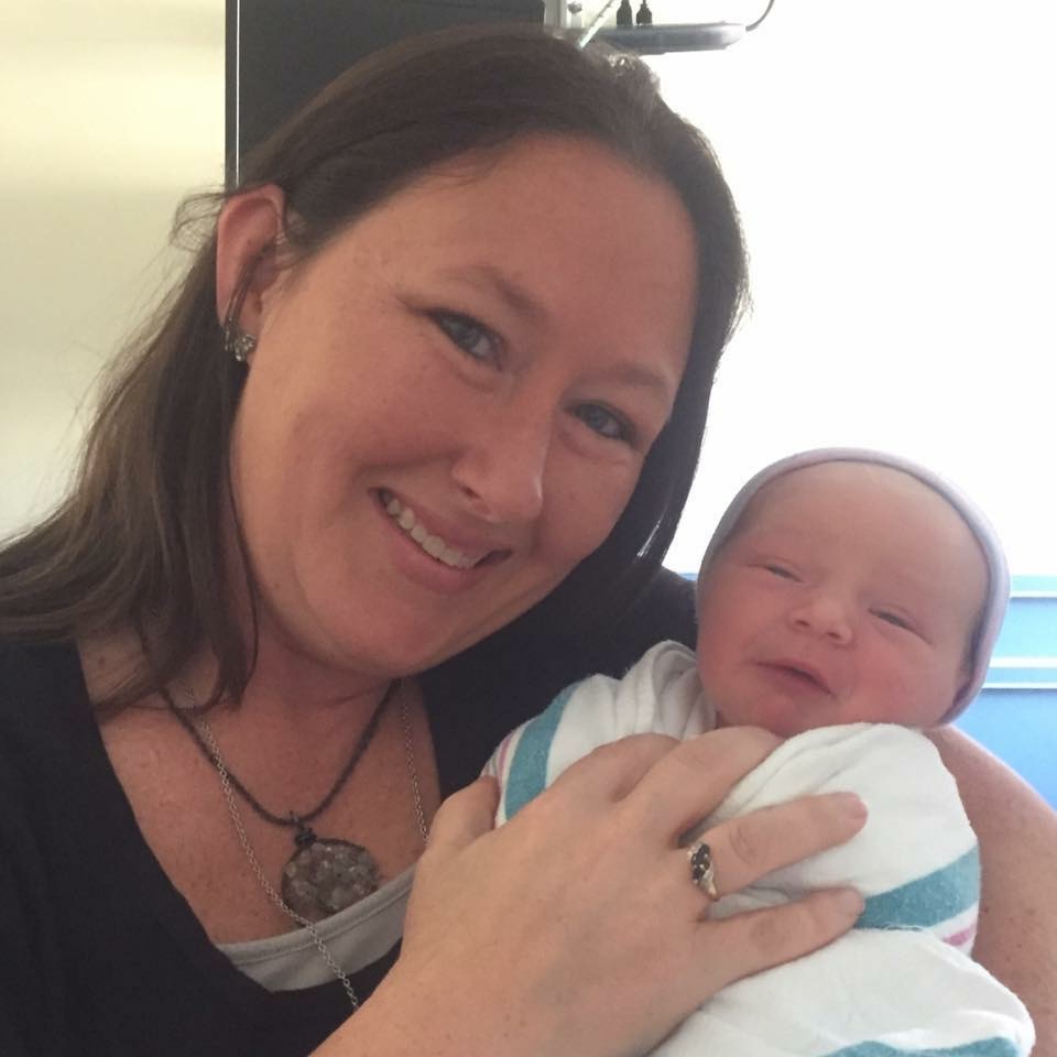 Kristie smiling, holding a newborn in a hospital blanket and hat