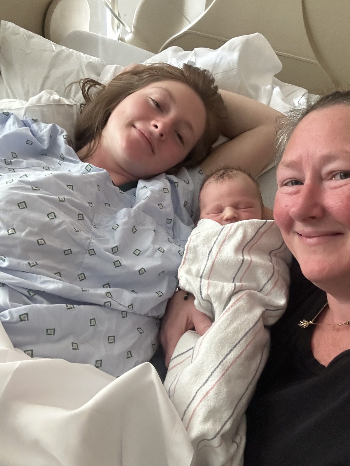 Kristie smiling beside a new mom resting in a hospital bed with her swaddled newborn